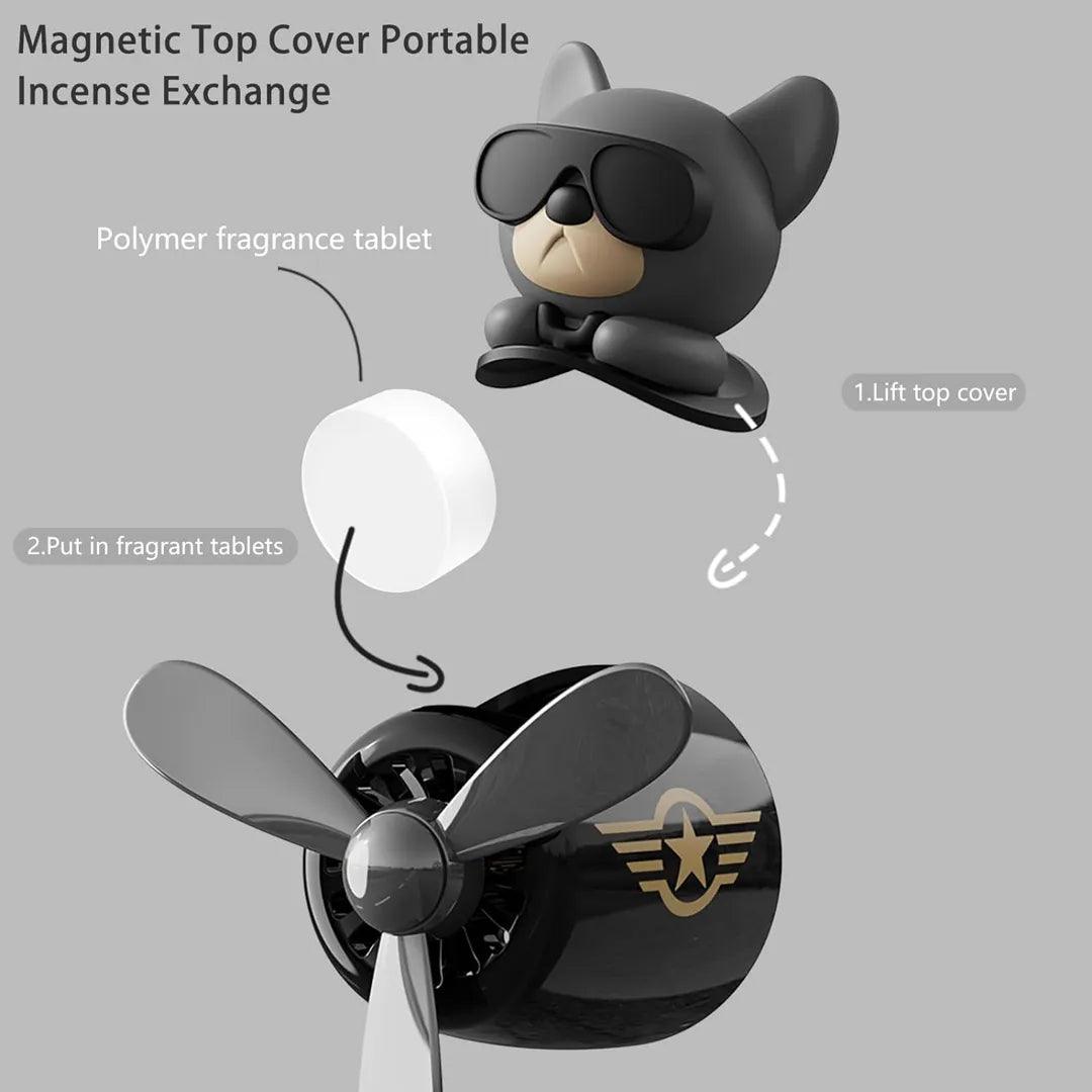 The image shows a car air freshener with a magnetic top cover for easy fragrance exchange. The design features a cute dog pilot wearing sunglasses. The process is demonstrated in two steps: 1) lift the top cover, and 2) insert the polymer fragrance tablet. The air freshener includes a propeller and a logo, emphasizing its portability and ease of use.