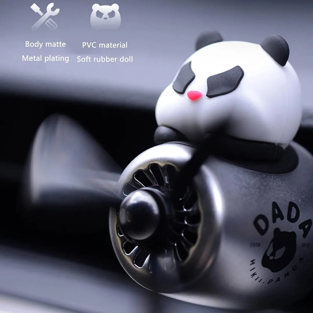 A panda-themed car air freshener with a black propeller, mounted on a silver and black airplane-shaped body. The panda figure sits on top with a pink nose and black eye patches. The text on the body reads 'DADA' with a panda logo and 'ESTD 2022'. Above the air freshener are icons and text indicating 'Body matte', 'Metal plating', 'PVC material', and 'Soft rubber doll'. The background shows a part of the car interior.
