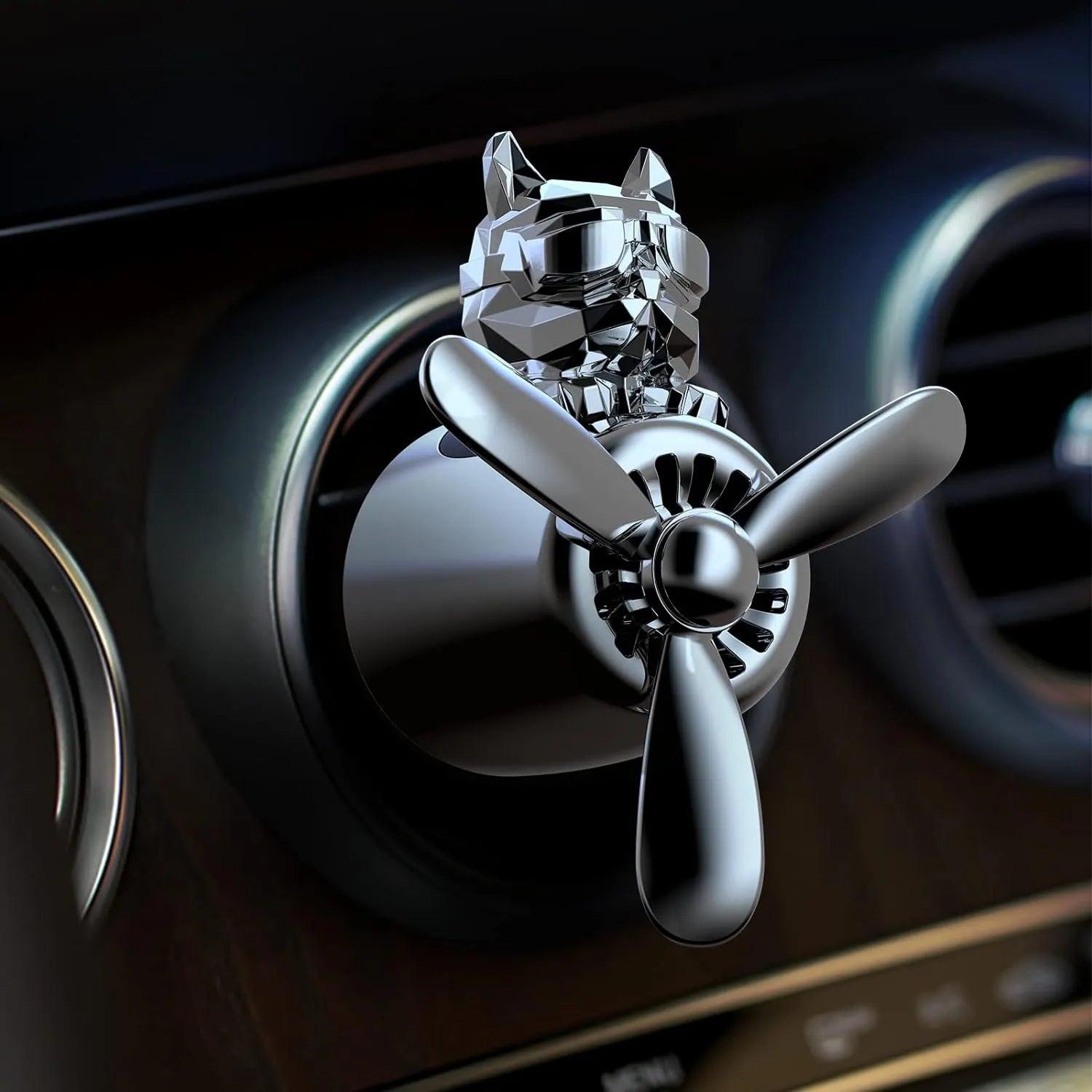 The image shows a close-up of a bulldog pilot car air freshener with a propeller design attached to a car's air vent. The bulldog figure has a sleek, faceted surface and is wearing aviator goggles. The air freshener has a metallic finish and the propeller is positioned at the front, indicating it is ready to disperse fragrance when the car's air conditioning or heating system is on. The background displays the car's dashboard and air vent controls.